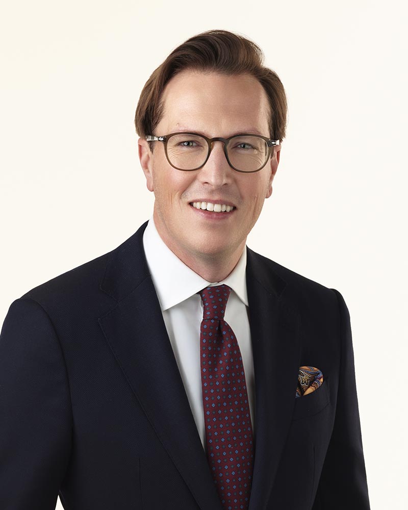 Caucasian man with eyeglasses in a dark suit with a red tie.